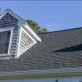 Dormer window with grey shingle siding and dark asphalt roof against a clear blue sky, showing a house exterior.
